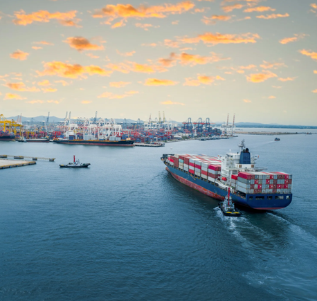 Aerial view of a cargo ship loaded with shipping containers approaching a commercial port with cranes, vessels, and container stacks in Australia.